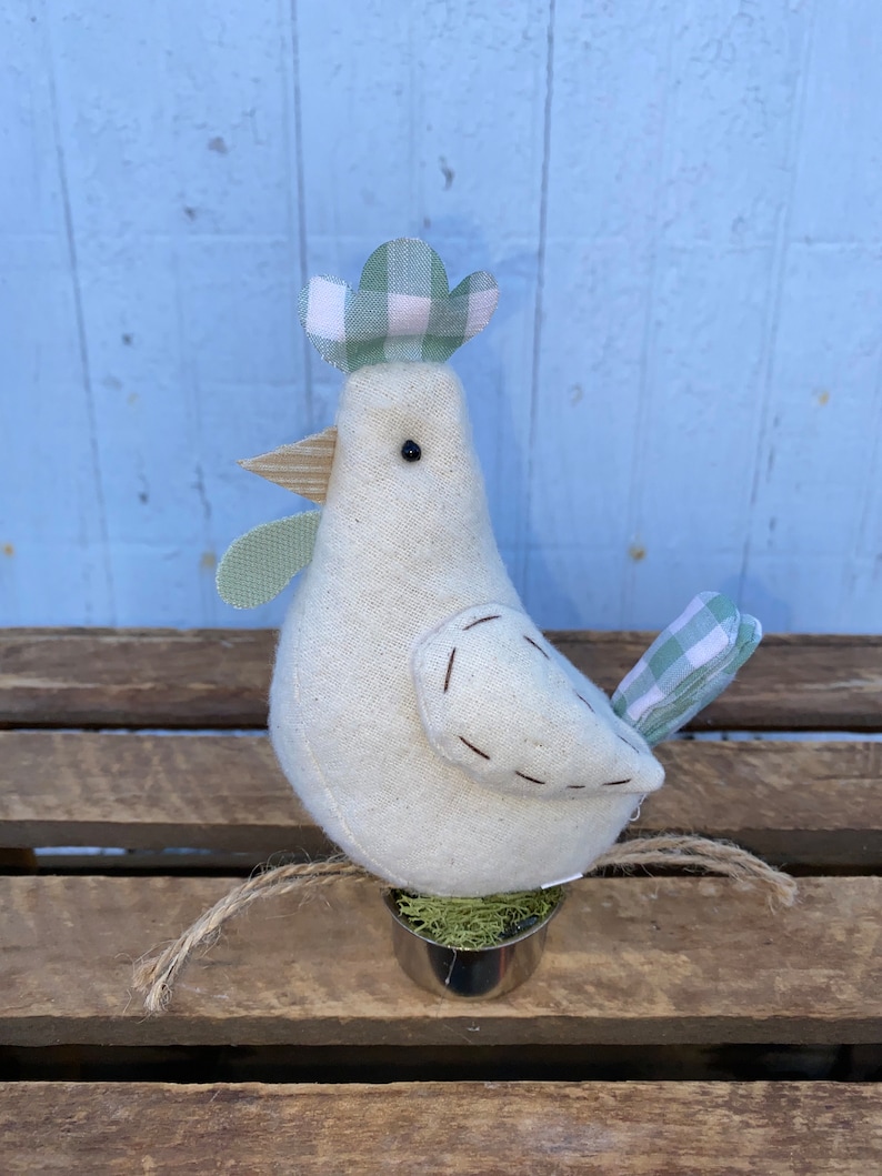 Puede incluir: Un pollo de tela blanca con un sombrero a cuadros verde y blanco, un pico marr&oacute;n y ojos negros. El pollo est&aacute; sentado en una maceta peque&ntilde;a con musgo verde.