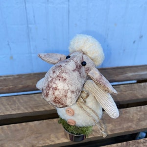 Puede incluir: Una peque&ntilde;a oveja de peluche blanca con manchas marrones y un gorro de lana blanco. La oveja est&aacute; de pie en una maceta peque&ntilde;a con musgo verde y una nariz de bot&oacute;n marr&oacute;n.