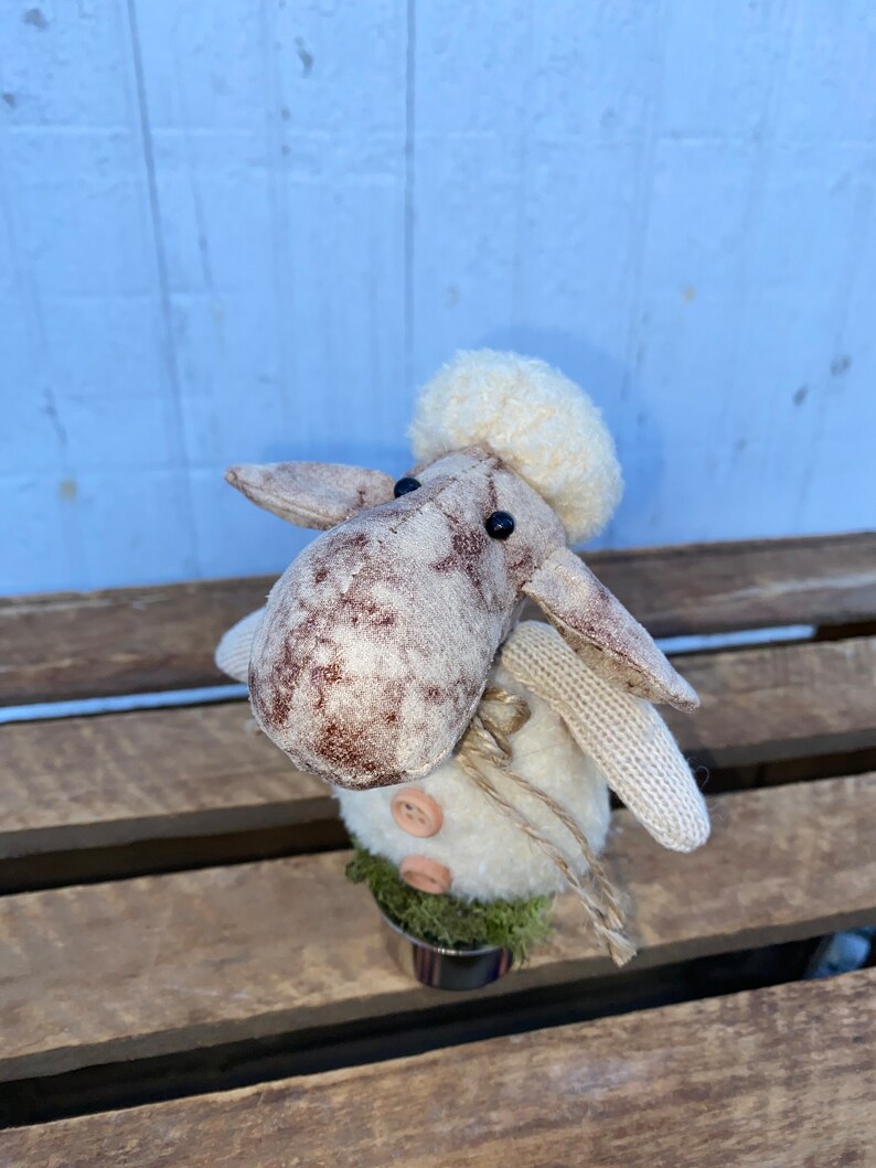 Puede incluir: Una peque&ntilde;a oveja de peluche blanca con manchas marrones y un gorro de lana blanco. La oveja est&aacute; de pie en una maceta peque&ntilde;a con musgo verde y una nariz de bot&oacute;n marr&oacute;n.