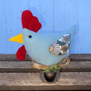Puede incluir: Un pollo de tela azul claro con una cresta roja, un pico amarillo y un ala con estampado floral. El pollo est&aacute; sentado en un nido de paja y musgo en una copa de plata.