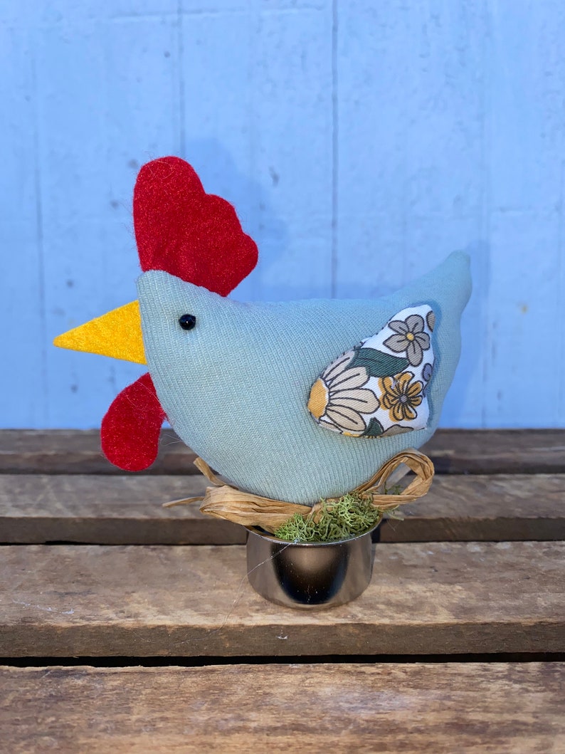 Puede incluir: Un pollo de tela azul claro con una cresta roja, un pico amarillo y un ala con estampado floral. El pollo est&aacute; sentado en un nido de paja y musgo en una copa de plata.