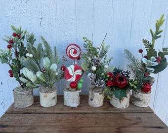 May include: Six small decorative arrangements in birch wood bases. The displays feature artificial greenery, pine needles, red berries, white berries, pinecones, and two round red and white peppermint candy ornaments on sticks.