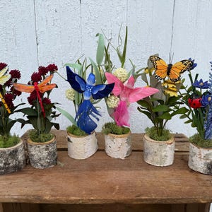 May include: A collection of decorative floral arrangements featuring various insects and creatures. Each arrangement is set in a birch bark pot and includes a dragonfly, butterfly, fairy, and ladybug. The color palette includes yellow, orange, blue, and pink.