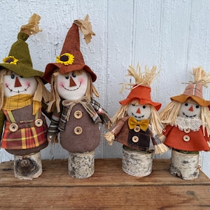May include: Four scarecrow dolls with fabric bodies, straw hair, and pointed hats, each mounted on a cylindrical birch wood base. The dolls feature stitched facial expressions, sunflower accents, and clothing in shades of brown, orange, and green.
