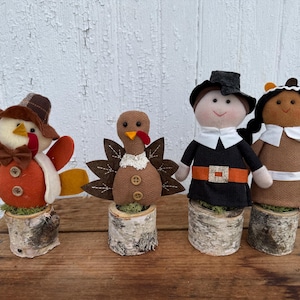 May include: Four decorative figures standing on birch wood log bases. Two figures are turkeys with orange and brown fabric bodies, and two are human figures wearing black and brown outfits with white collars, all featuring button accents.