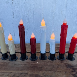 May include: A collection of seven battery-operated LED taper candles in varying heights and colors. The candles are cream, white, and red, with realistic flickering flame tips. Each candle sits in a black metal base, creating a warm, inviting ambiance.
