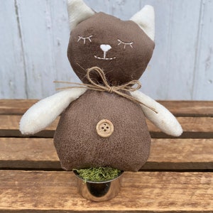 Puede incluir: Un gato de peluche marr&oacute;n con orejas blancas y una nariz de bot&oacute;n, sentado en una peque&ntilde;a maceta de metal con musgo verde. El gato tiene ojos bordados y una sonrisa.