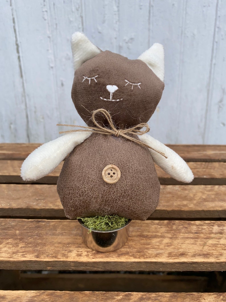 Puede incluir: Un gato de peluche marr&oacute;n con orejas blancas y una nariz de bot&oacute;n, sentado en una peque&ntilde;a maceta de metal con musgo verde. El gato tiene ojos bordados y una sonrisa.