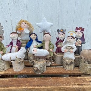 May include: A felt nativity scene with figures of Mary, Joseph, baby Jesus, a shepherd, an angel, three wise men, a sheep, and a camel. The figures are standing on birch wood logs.