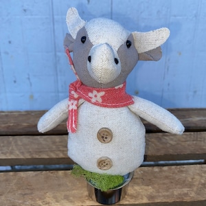 Puede incluir: Un juguete de peluche de vaca blanco y gris que lleva una bufanda roja y blanca con flores blancas. El juguete est&aacute; sentado en una maceta peque&ntilde;a con musgo verde.