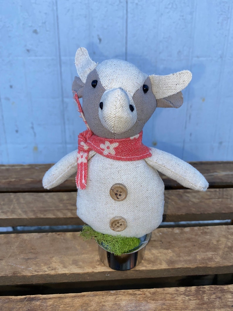 Puede incluir: Un juguete de peluche de vaca blanco y gris que lleva una bufanda roja y blanca con flores blancas. El juguete est&aacute; sentado en una maceta peque&ntilde;a con musgo verde.