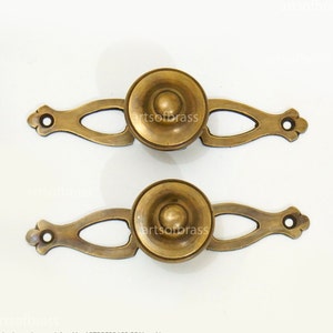 May include: Two brass drawer pulls with a decorative design. Each pull has a round knob with a raised centre and a long, curved handle with an openwork design.