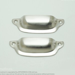 Puede incluir: Dos tiradores de cajón de armario plateados con forma rectangular redondeada. Los tiradores tienen un acabado liso y pulido.