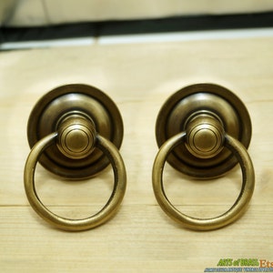 May include: Two antique brass cabinet door pulls with a ring handle. The pulls have a round base with a raised centre and a decorative ring handle.