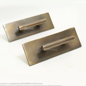 May include: Two antique brass cabinet pulls with a rectangular base and a cylindrical handle. The pulls have a distressed finish.
