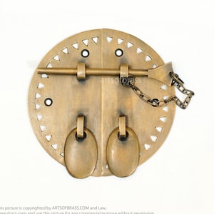 May include: A brass door latch with a circular design and two oval handles. The latch has a decorative chain and a metal bar with a pointed end.