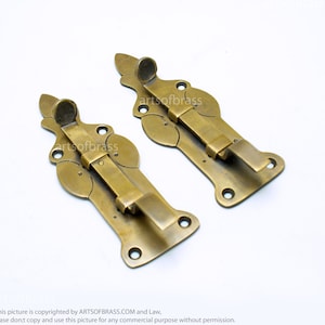 May include: Two antique brass door latches with ornate designs. The latches have a decorative, floral pattern and a sliding bolt.