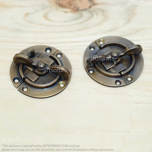 May include: Two antique brass cabinet door catches with a round shape and a latch mechanism. The catches are on a wooden surface.