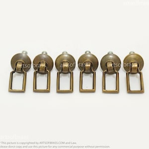 Peut inclure: Six boutons de cabinet en laiton antique de forme carrée avec une petite poignée en boucle. Les boutons ont une finition légèrement vieillie.