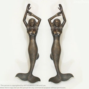 May include: Two bronze-colored mermaid door handles. Each handle features a mermaid with arms raised, a scaled tail, and a handle at the base. The text "artsofbrass" is visible in the image.