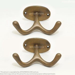 Könnte beinhalten: Zwei antike Doppelhaken aus Messing mit ovalen Rückplatten. Jeder Haken hat ein einzigartiges, geschwungenes Design. Die Rückplatten tragen den gravierten Text "artsofbrass".