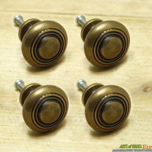 Pode incluir: Quatro botões de armário de latão antigo com um design circular e um centro elevado. Os botões têm um acabamento ligeiramente envelhecido e desbotado.