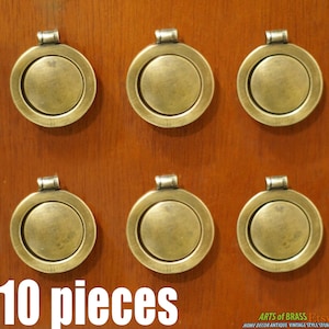 Puede incluir: Diez pomos redondos de armario de latón antiguo con un anillo decorativo alrededor del borde. Los pomos se muestran en una superficie de madera.
