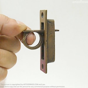 May include: Brass door latch with a rectangular shape and a ring handle. The latch has a red accent on the bottom.
