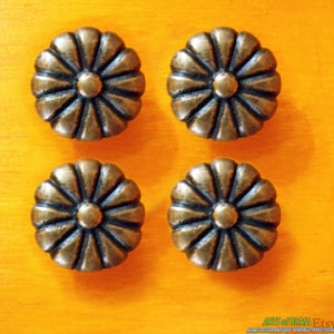 May include: Four antique brass flower-shaped cabinet knobs. Each knob has a detailed petal design and a central rounded accent. The knobs are arranged on a bright yellow-orange surface, creating a striking contrast.