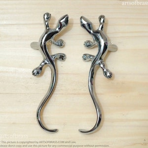 May include: Two silver lizard-shaped door handles with a curved tail. The handles are on a wooden surface.