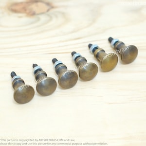 Könnte beinhalten: Sieben antike Messing-Schrankknöpfe mit runder Oberseite und einer Gewindeschraube. Die Knöpfe sind in einer Reihe auf einer hellbraunen Holzoberfläche angeordnet.