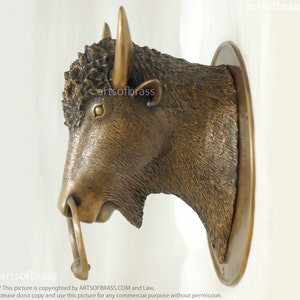 May include: A bronze bison head wall mount with a ring on the nose. The bison is facing to the right and has a detailed, textured surface.