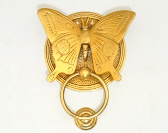 Puede incluir: Un llamador de puerta dorado en forma de mariposa con un anillo redondo. La mariposa tiene alas detalladas y un cuerpo texturizado.