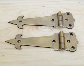 May include: Two antique brass hinges with a decorative fleur-de-lis design. The hinges are closed and have a polished finish.