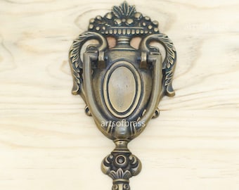 Könnte beinhalten: Ein dekorativer Türklopfer aus Messing in Form einer Vase mit floralem Design. Der Türklopfer hat einen Ringgriff und eine detaillierte Basis.