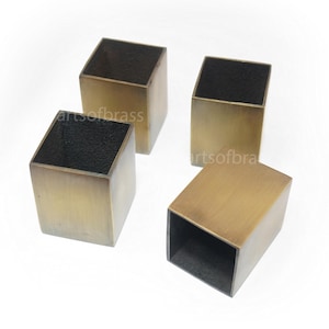 May include: Four square-shaped containers made of brushed brass metal. Each container features a smooth, metallic exterior finish with a dark, textured interior. The items are arranged in a cluster, with three standing upright and one lying on its side.
