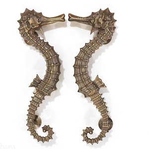 Può includere: Un paio di maniglie per porte in ottone ornate a forma di cavallucci marini. Le maniglie sono rivolte l'una verso l'altra e presentano un design dettagliato e testurizzato.