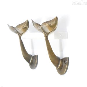 May include: Two brass whale tail hooks mounted on a white shelf. The hooks are in the shape of a whale tail and are a decorative and functional addition to any home.