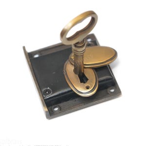 Peut inclure: Un mécanisme de serrure carré en métal noir avec une clé en laiton insérée dans une plaque d'entrée de serrure ovale en laiton. La clé possède un anneau ovale et une tige cylindrique. La plaque comporte quatre trous de vis aux coins.