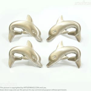 Könnte beinhalten: Vier silberfarbene dekorative Objekte in Delfinform. Jeder Delfin hat einen gebogenen Körper, eine Rückenflosse und eine Schwanzflosse. Die Delfine sind auf weißem Hintergrund angeordnet. Der Text "artsofbrass" ist sichtbar.