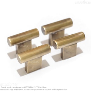 May include: Four antique brass cabinet knobs with a rectangular base and a rounded top. The knobs are arranged in a staggered pattern on a white surface.
