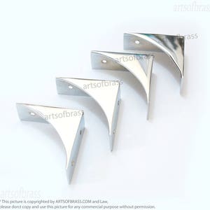 May include: Four silver metal corner brackets, each with a curved design, arranged in a row on a white background.