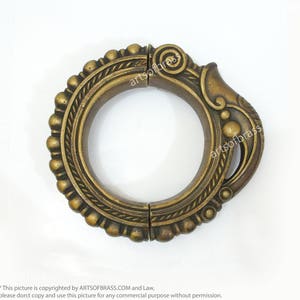 May include: A gold-toned metal bangle bracelet with intricate detailing and a decorative design. The bracelet is hinged and opens to fit the wrist.