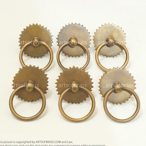 Può includere: Sei pomelli per armadi in ottone con un bordo circolare a festoni e un tiretto ad anello. I pomelli sono di un colore ottone antico caldo.