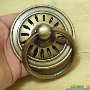 May include: A brass ring pull cabinet door handle with a decorative, circular, openwork design. The handle is in a hand, showing the size and detail.