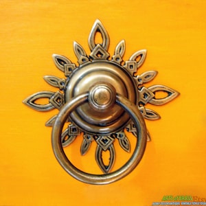 May include: A brass door pull with a sunburst design. The pull has a ring handle and a decorative center piece.