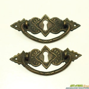 以下が含まれることがあります： 鍵穴のデザインが施されたアンティークな真鍮製の引き出しノブ2個。ノブは、中央にダイヤモンドの形をした装飾的な華やかなデザインが施されています。