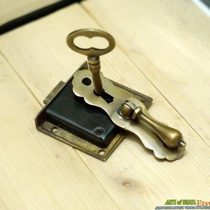May include: Antique brass lock with a key and a decorative handle. The lock has a black metal plate and is mounted on a wooden surface.