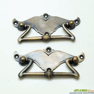 May include: Two antique brass drawer pulls with a decorative, ornate design. The pulls have a curved, rectangular shape with a centre bar.