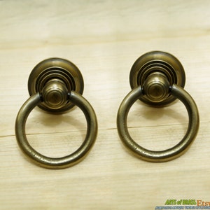 May include: Two antique brass ring pulls with a decorative design. The pulls have a circular ring and a round base with concentric circles.
